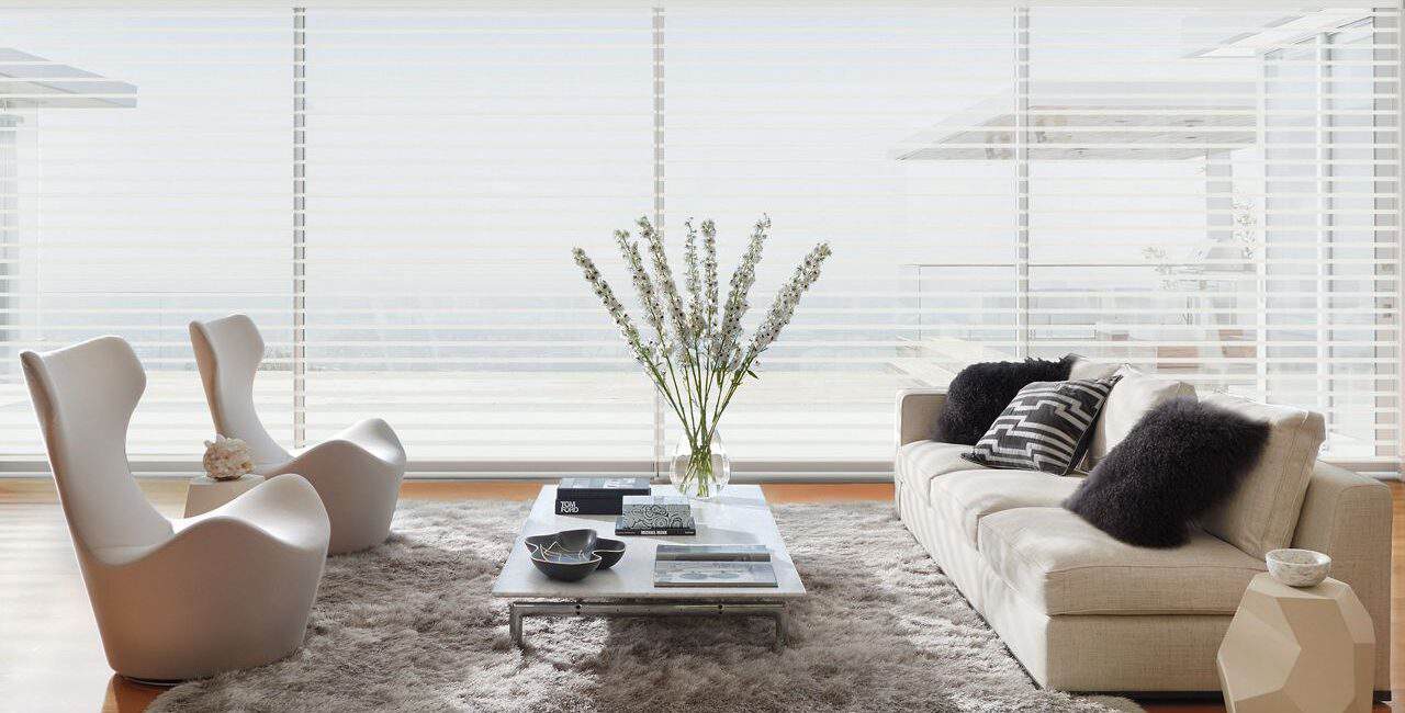 Bright living room with beige sofa, white chairs, and glass coffee table in front of floor-to-ceiling window blinds.