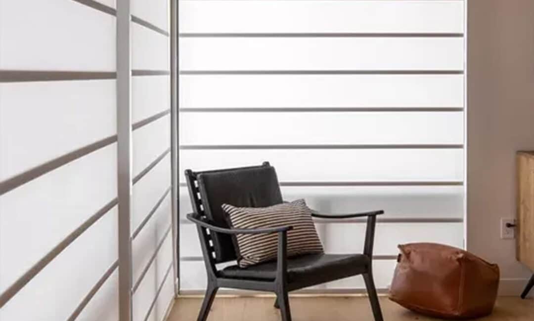 Stylish black lounge chair with striped pillow placed in a well-lit modern room featuring large white window blinds.