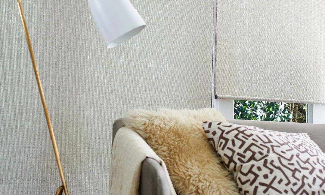 A comfortable living room corner featuring a beige sofa with decorative pillows, a cozy blanket, and a stylish floor lamp against a window with natural light and green views.