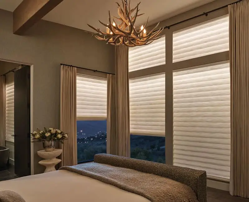 Elegant bedroom with large windows featuring white pleated shades and cream curtains, creating a warm and inviting atmosphere with soft lighting.