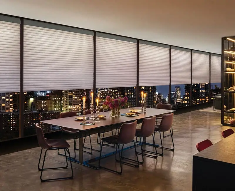Stylish dining area with city views, white roller blinds, contemporary table, pink flowers, candles, and cityscape at night, ideal for sophisticated gatherings.