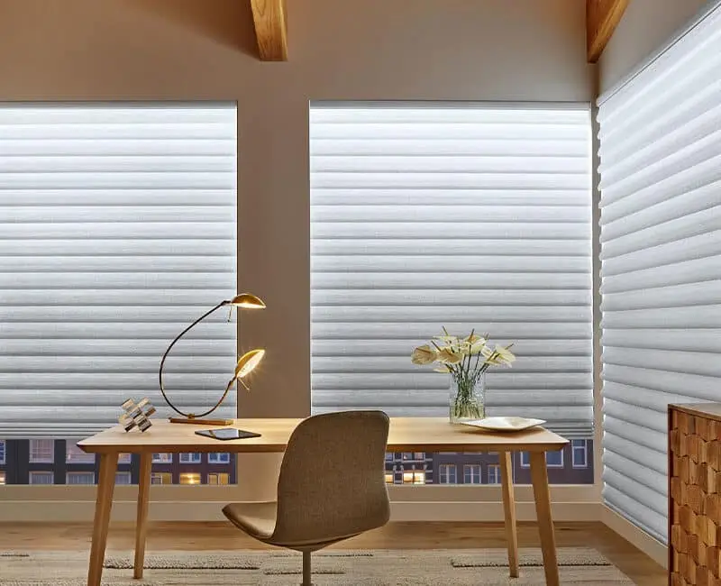 Bright workspace with blinds, wooden desk, and comfortable chair. Elegant lamp, flower vase, and city view create a peaceful work environment. Perfect for home or office use.