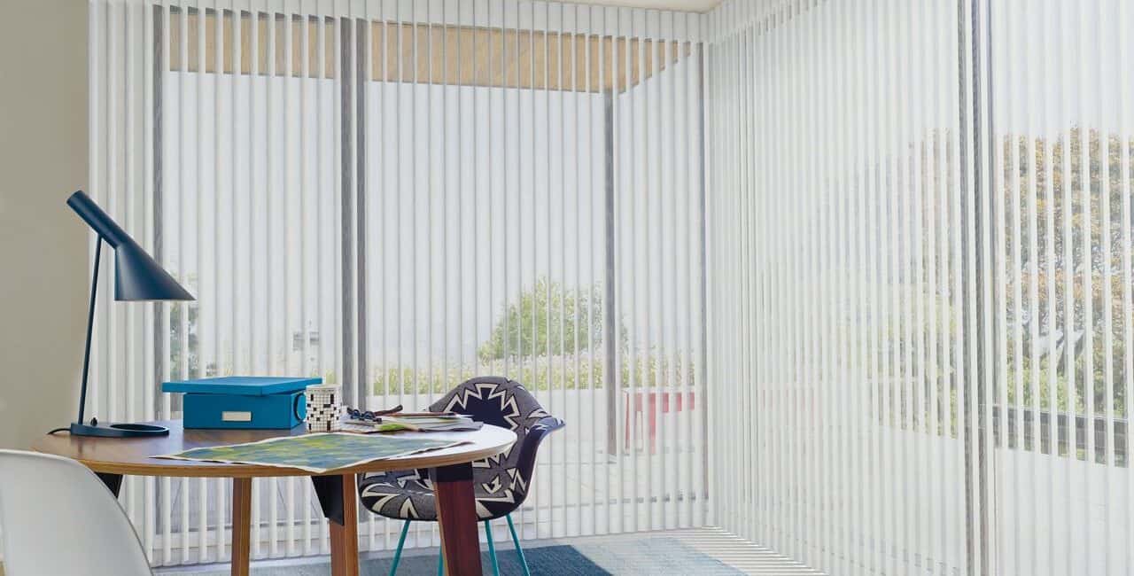 Vertical blinds in a modern bright room with large windows, providing privacy and light control. Minimalist decor with a small table, chair, and outdoor view.