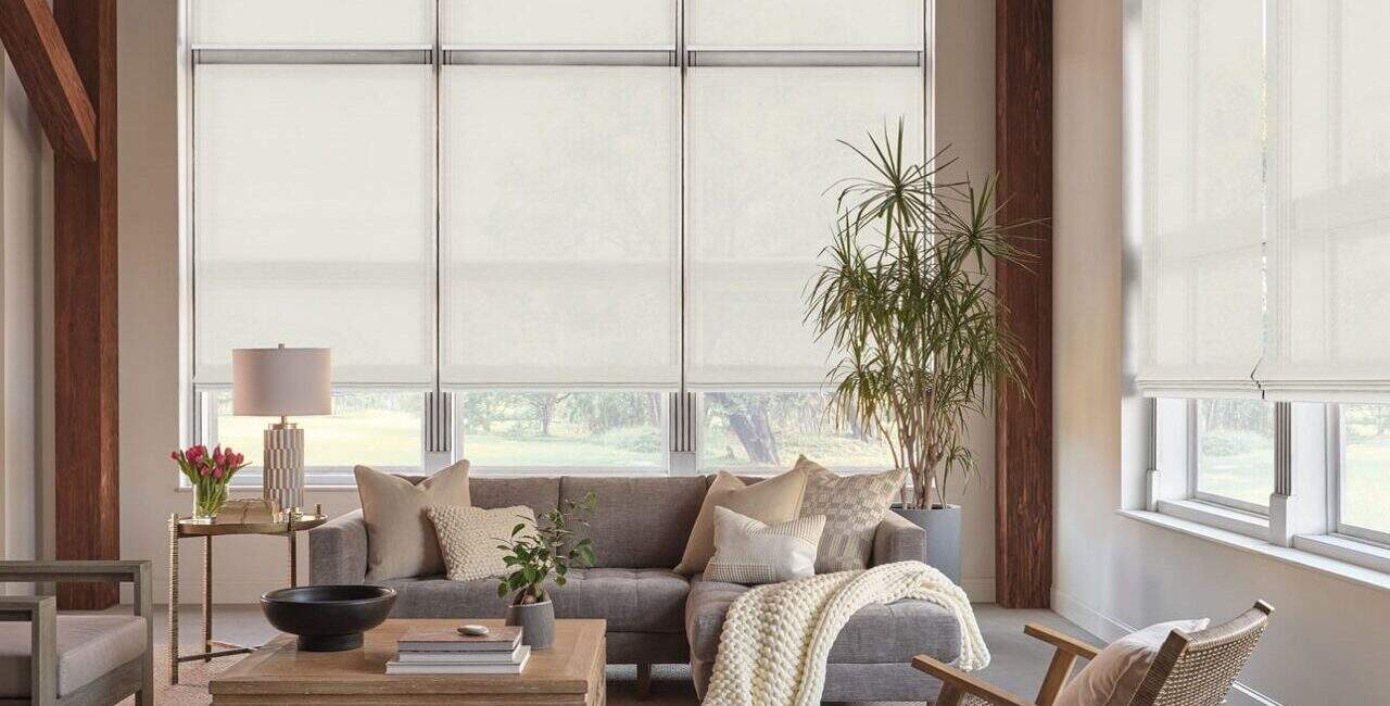 Bright living room with large windows featuring roller blinds, cozy sofa, armchair, potted plants, and neutral tones, providing a warm and inviting atmosphere.