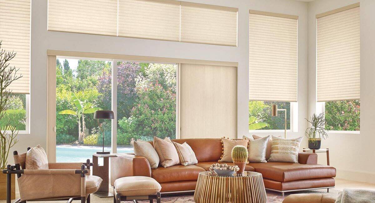 Spacious living room featuring large windows with beige blinds, a tan leather sofa, contemporary decor, and views of greenery outside. Bright, airy, and stylish for relaxing or entertaining.