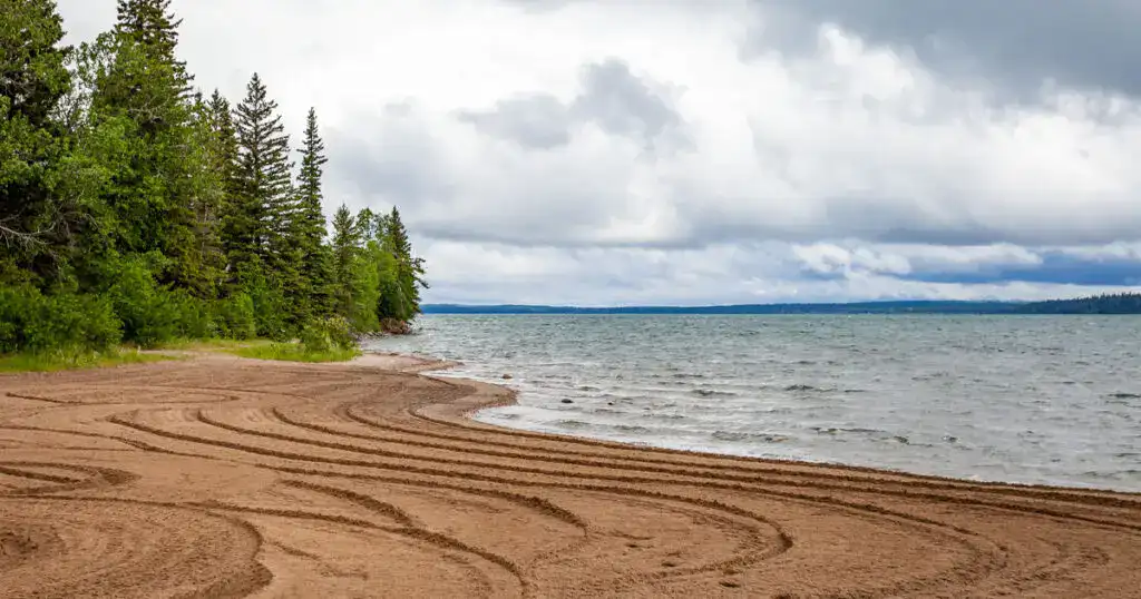 Scenic view of Clear Lake in Manitoba featuring a sandy beach, calm waters, and lush green trees under a cloudy sky. Perfect for outdoor recreation and nature photography.