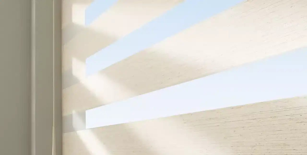 Bright sunlight filters through white horizontal window blinds, creating patterns of light and shadow, with a view of lush greenery outside.