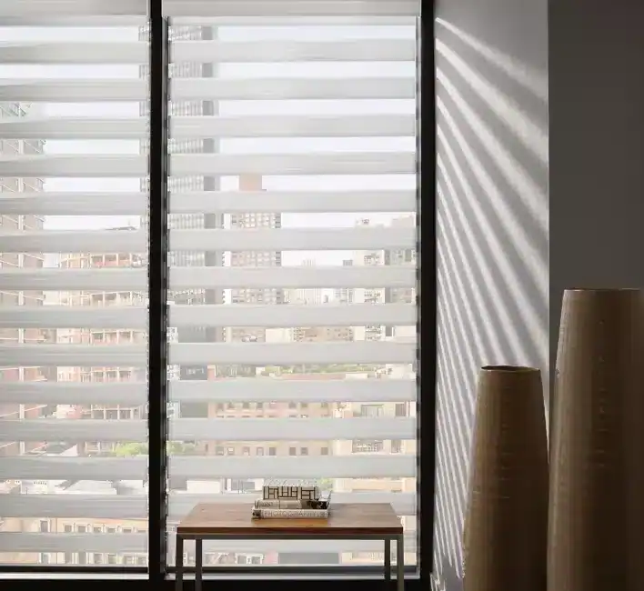 Modern apartment window with horizontal blinds casting light and shadows, city skyline visible outside.
