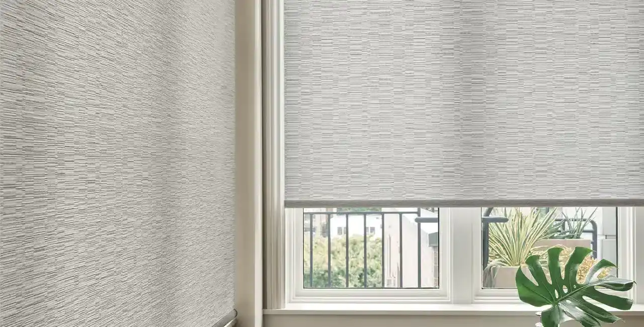 ALT text: Interior window scene with textured roller blinds, a small table with decorative vases and a Monstera leaf, framed by a classic white window and wall trim.