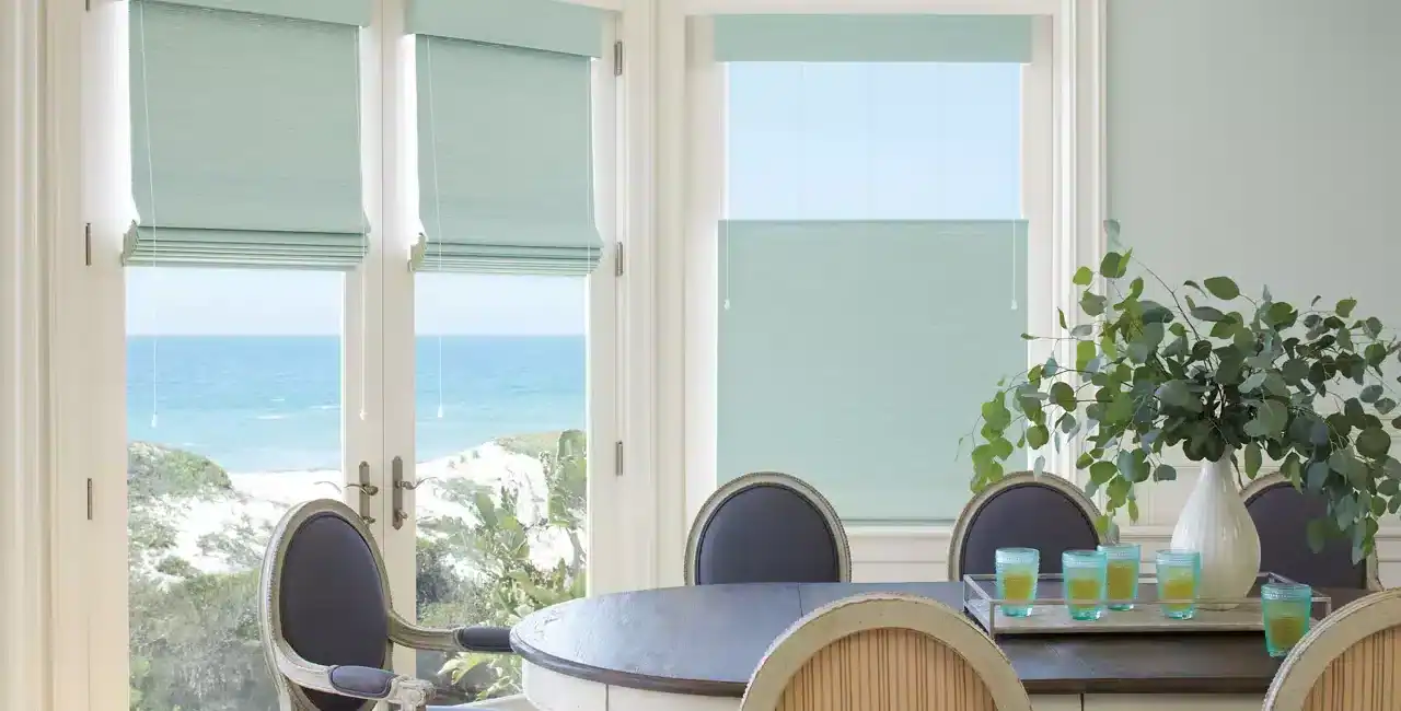 Bright seaside dining area featuring large windows overlooking the ocean with blue shades and greenery, elegant furniture, and table settings for a relaxing coastal meal.