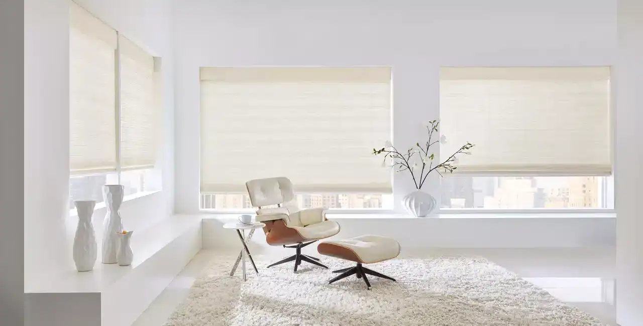 Bright and airy interior with large windows featuring cream roller blinds, modern beige lounge chair with matching ottoman, plush white rug, and elegant white vases with minimal decor.