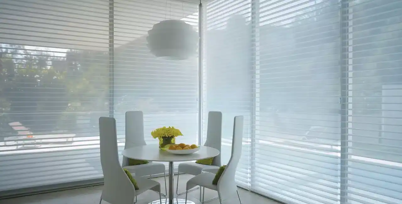 Bright, airy breakfast nook with large windows, white chairs, and a round table decorated with flowers and fruit.