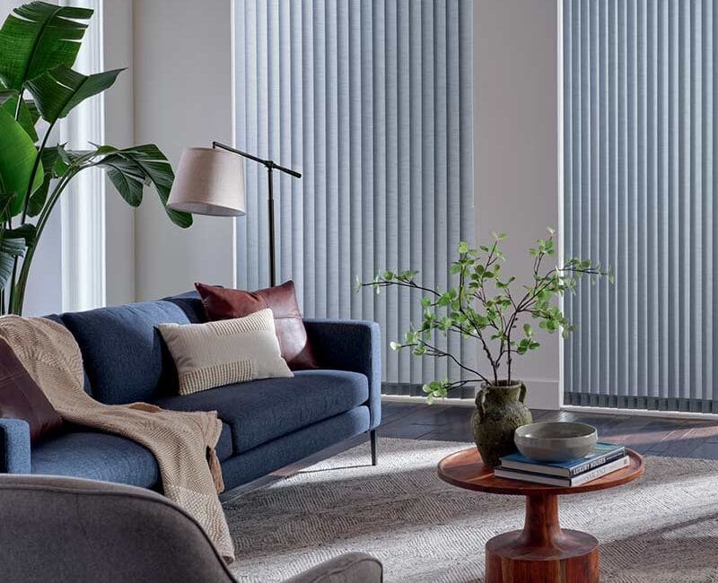 Bright living room with large windows, vertical blinds, lush green plants, modern sofa, and minimalist wooden coffee table, creating a stylish and relaxing space.
