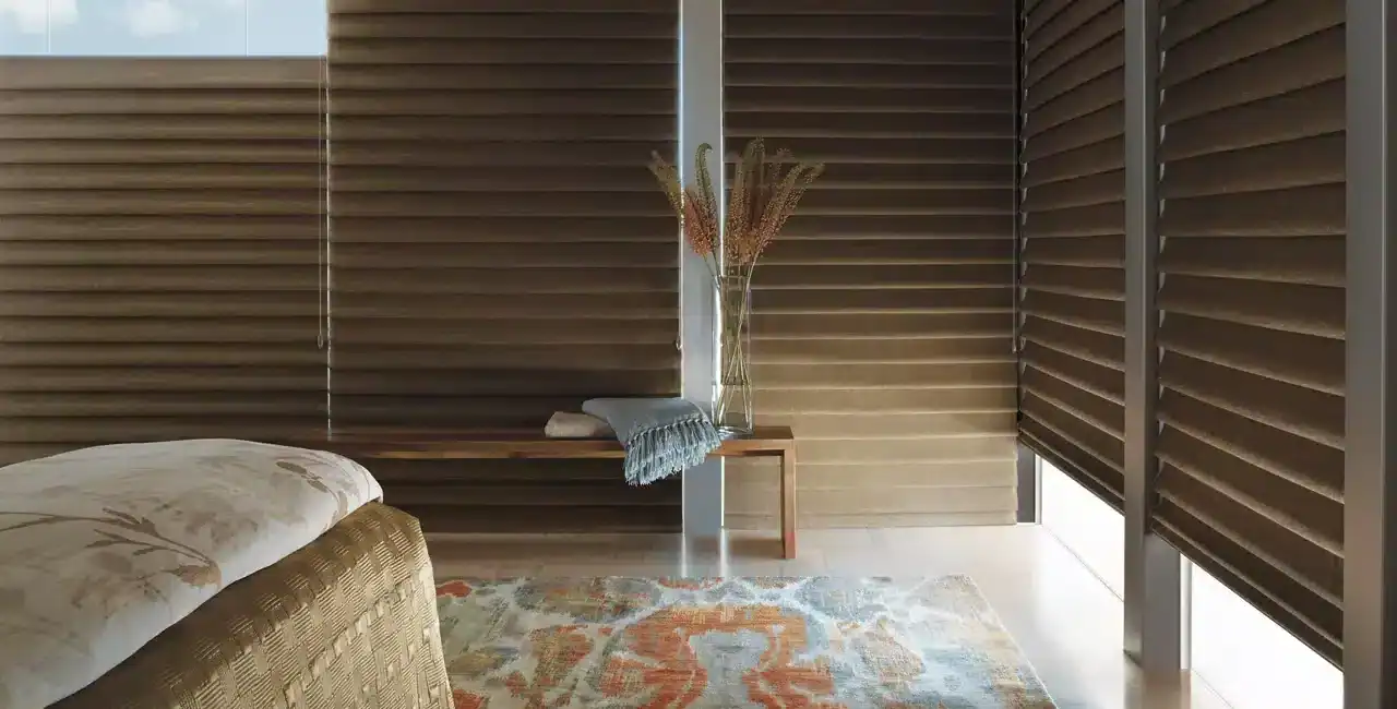 Close-up of a bedroom with brown horizontal window blinds, a bed with floral bedding, a wooden bench with a vase of dried flowers, and a colorful rug creating a warm, modern ambiance.